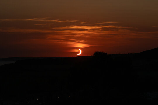 Son Bou, Spain - 12 August 2026: View of a partial solar eclipse at sunset with the sun partially obscured by the moon, casting a warm orange glow over the dark horizon.