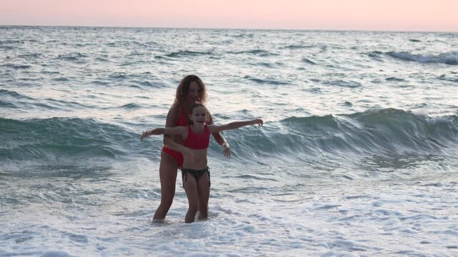 Coastal vacation fun with smiling mother and daughter in red swimwear enjoying sea surf at sunset. Medium tracking shot.