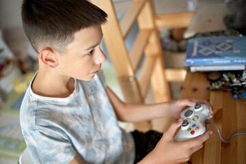 Young boy plays a video game at a wooden desk, holding a wired controller in both hands. Warm indoor light and a cozy bedroom workspace create a focused childhood leisure scene. © Yulia Mihaylova
