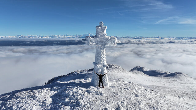 Geneva, Switzerland - 12 August 2026: View of a person standing with arms raised in front of a snow-covered cross on a mountain peak above a sea of clouds under a clear blue sky.