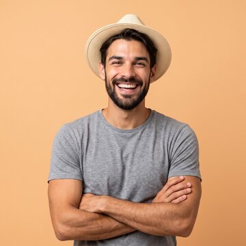 A handsome young man with a beard and a straw fedora hat, smiling confidently with his arms crossed while standing against a solid beige studio background.
