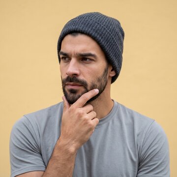A contemplative young man with a beard and dark grey beanie looking away against a soft yellow wall