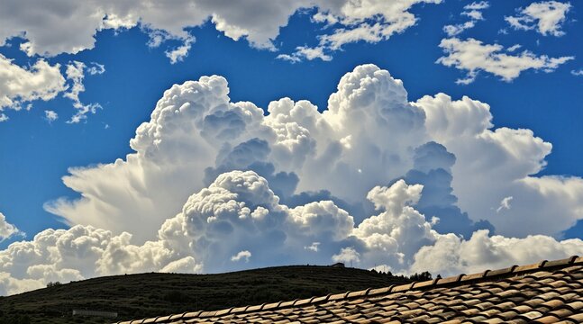 Large fluffy white clouds drift over green grassy hill landscape under deep blue summer sky. Bright daylight illuminates rolling terrain with natural sunlight. Scenic rural view features textured