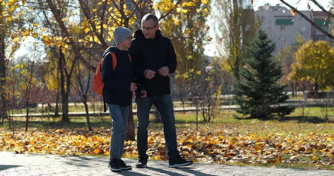 Father and son walk together in autumn park. Dad and child enjoy conversation while surrounded by colorful fall leaves. Warm atmosphere captures family bonding outdoors.