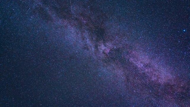 Vivid astrophotography timelapse of the Milky Way and countless stars moving across the clear night sky, showing a dense celestial field in glowing blue and purple tones with faint satellite trails.