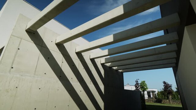 Concrete architecture with a modern pergola design casting shadows on a brutalist wall, revealing a minimalist home facade and a clear blue sky on a sunny summer day