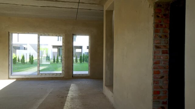 Sun moving inside unfinished room with plastered walls and concrete floor during new modern house construction, revealing bright backyard with green grass and trees