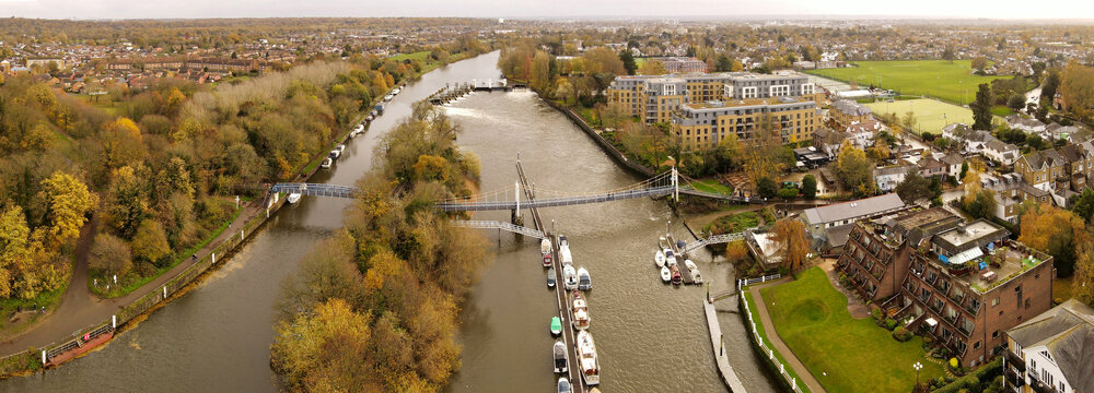 Aerial view of Teddington Lock on the River Thames with moored boats, a pedestrian footbridge, and surrounding autumn foliage in Teddington, England, United Kingdom.