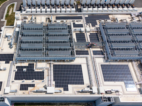 Aerial view of a modern hyperscale data center featuring extensive rooftop solar panel arrays, industrial cooling units, and complex infrastructure in Melegnano, Lombardy, Italy.