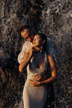Sensual couple standing under waterfall spray, intimate lovers enjoying sunset light