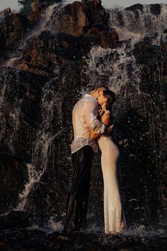 Passionate romantic couple kissing and hugging under waterfall near dark wet rocks at sunset