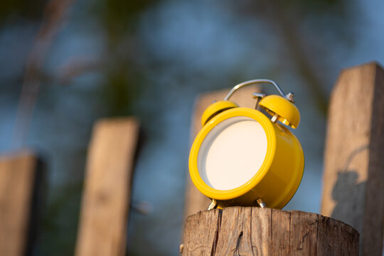 A bright yellow retro alarm clock is standing on an old wooden fence. Instead of a dial, there&rsquo;s an empty space for text.
