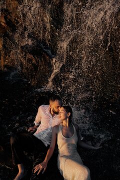Passionate couple lying on wet rocks under waterfall spray during sunset