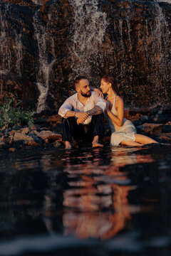 Romantic couple sitting near waterfall with water reflection, young man and woman looking at each other at sunset