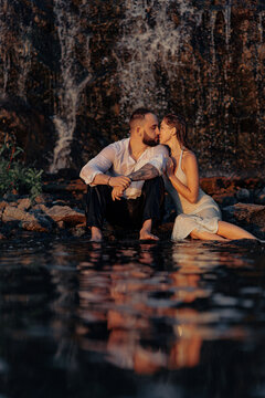 Passionate couple kissing sitting near waterfall with water reflection, romantic lovers on rocky shore at sunset