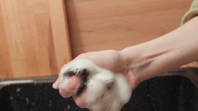Close-up of hand gripping wet black sponge covered in thick white foam over dark kitchen sink, showing visible pressure, dripping moisture, suds, texture, and practical household cleaning task