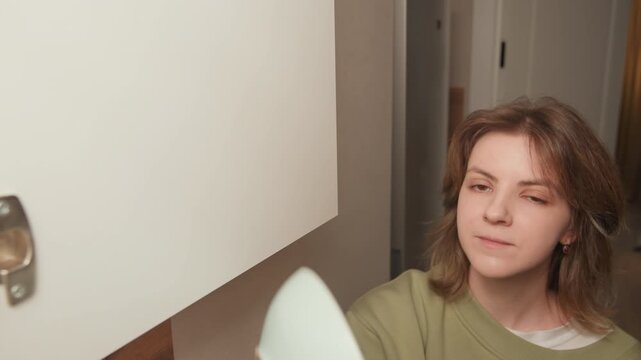 Young woman leans forward and reaches toward white cabinet or mirror, arm extended with calm focused expression, creating subtle sense of interaction inside cozy interior space, with woman, reaching