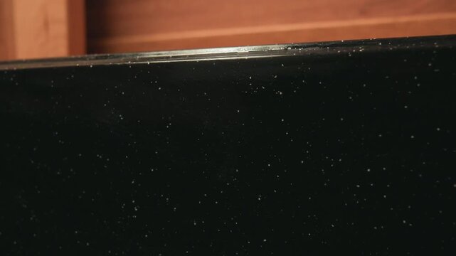 Close-up frontal view shows hand pressing red cloth across dark reflective panel, creating downward wiping motion while tiny paint chips, specks, and surface marks remain visible, with wipe, clean