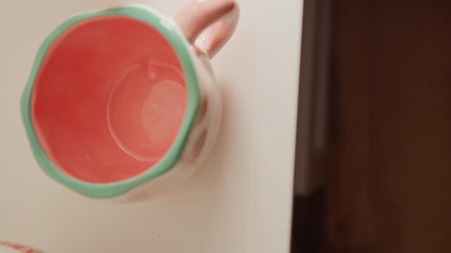 Close-up view shows hand tilting ceramic mug to reveal empty coral-red interior and mint-green rim above white tabletop beside dark doorway, with gentle motion adding casual household context and