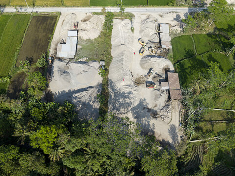 Aerial view of a stone crusher facility with piles of sand and gravel, heavy machinery, and trucks surrounded by lush green rice fields in Kecamatan Karangnongko, Jawa Tengah, Indonesia.