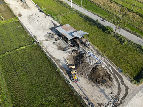 Aerial view of a stone crusher facility operating near green rice fields with piles of gravel and heavy machinery in Kecamatan Karangnongko, Jawa Tengah, Indonesia.
