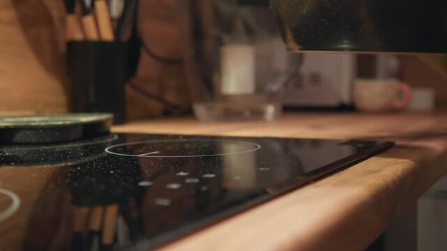 Close-up shows hand guiding speckled ceramic saucepan onto sleek black induction cooktop, with warm kitchen light, soft reflections, and shallow focus emphasizing cookware, surface, and motion