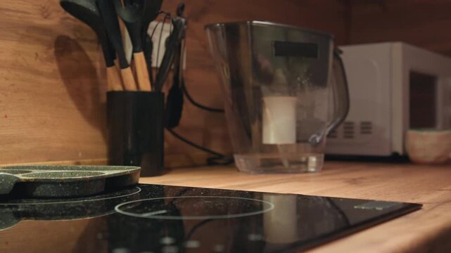 Close-up of hand gripping handle to lift small pot off glossy black cooktop, showing upward motion during meal preparation inside warm wooden kitchen with countertop, utensils, and soft natural light