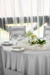 Fototapeta premium Elegant wedding reception table setting with white tablecloth, delicate floral centerpiece, crystal wine glasses, and plates against sheer curtains