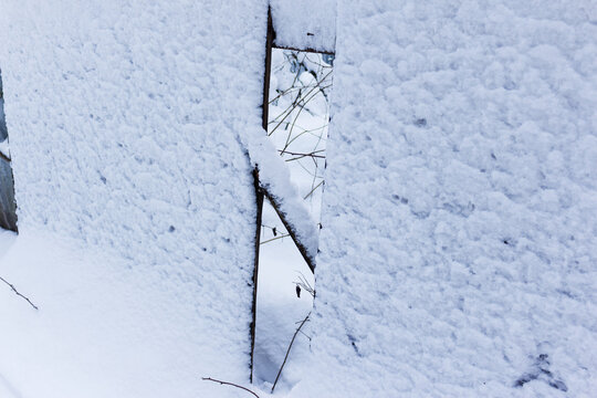 Broken wooden fence covered in thick white snow with a vertical gap, revealing glimpses of the frozen winter landscape behind during a cold overcast day in the countryside