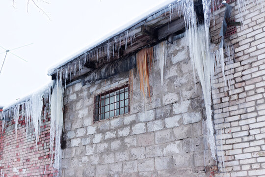 Long sharp icicles hanging from the roof edge of an old rustic brick building during a cold winter day, showcasing frozen water structures against a weathered facade
