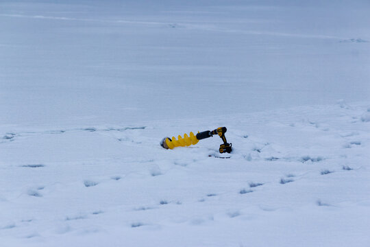 Yellow cordless ice auger resting on a vast snowy river surface, professional power tool for winter fishing prepared to drill holes in the thick frozen ice cover outdoors