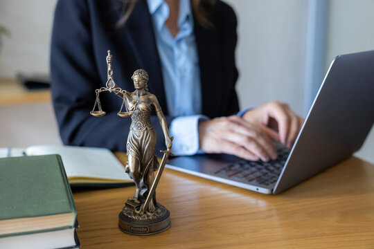 Lawyer in dark suit typing on silver laptop. Bronze Lady Justice figurine stands on wooden desk. Open notebook and pen nearby. Modern digital legal practice with traditional justice symbol in office.