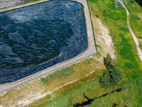 Aerial view of a sewage sludge storage facility with a large black membrane cover for odor control, surrounded by grassy embankments and a dirt path, Riga, Latvia.