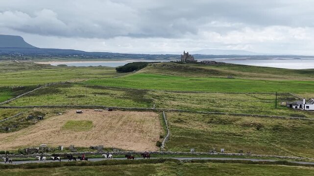 Aerial drone flight tracking forward over coastal fields towards Classiebawn Castle, with horse riders and horses on a path in the foreground, County Sligo, Ireland.