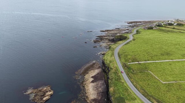 Aerial view flying forward along Mullaghmore Head coast, capturing sea kayakers in the water and a red car driving on the coastal road, Sligo, Ireland.