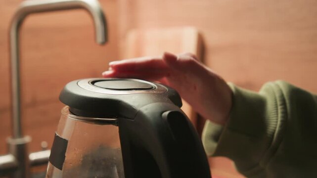 Right hand steadies black kettle lid while clear glass carafe fills with hot water on wooden kitchen counter, with steam, droplets, warm light, and domestic routine in view, with kettle, lid, hand