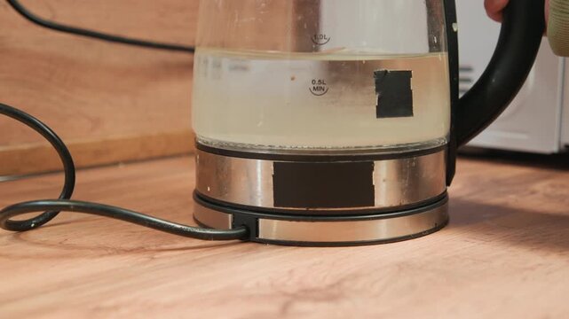 Transparent electric kettle sits on round hotplate atop wooden countertop with braided cord connected, water bubbling inside, steam rising and warm light reflecting across glass and metal surfaces
