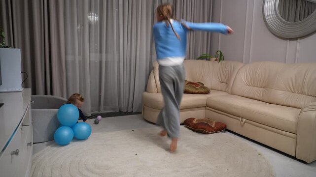 Happy young girl spinning and dancing in modern living room while her toddler sibling plays in ball pit nearby