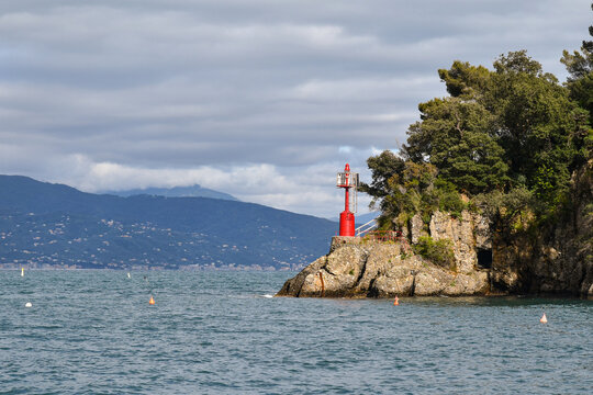 Small red maritime beacon lighthouse on the rocky cliffs of Punta del Coppo with green pine trees, Portofino, Liguria, Italy