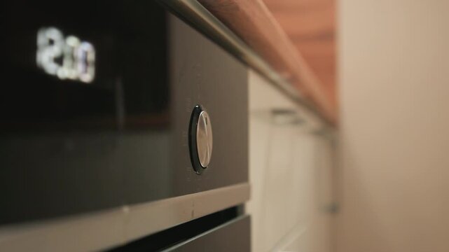 Hand reaches forward to turn black reflective oven control knob, visibly changing setting while partial digital display shows number; shallow focus keeps knob and hand sharp, background softly blurred