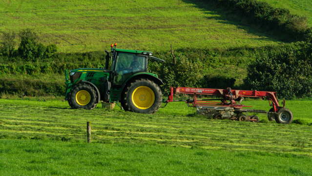 West Cork, Ireland, August 25, 2023. Green tractor with a red hay tedder on a vibrant, lush Meadow. Farming scene.