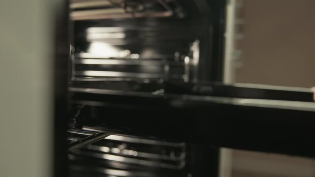 Person hand pulling metal oven rack outward inside empty oven, showing smooth kitchen appliance movement with reflective chrome shelves, polished stainless interior, warm neutral indoor light, and