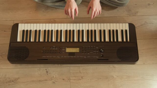 High overhead view of seated person with hands hovering above portable electronic keyboard on wooden floor, poised for playing with fingers ready above cream and black keys in soft indoor light