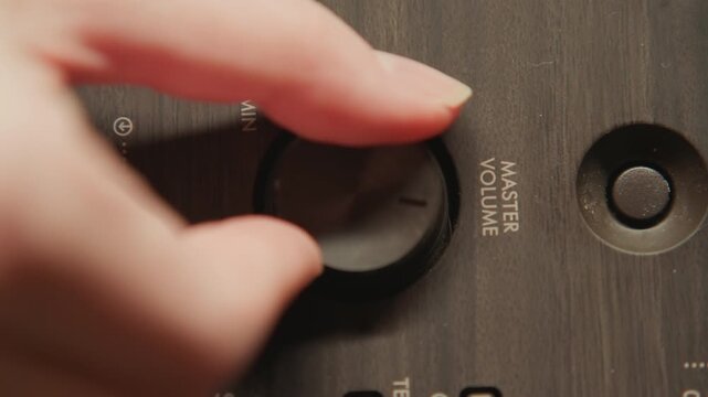 Close-up view of human hand turning brushed metal volume knob on audio amplifier control panel; knob positioned near center between engraved MIN and MAX markings and smaller recessed power button