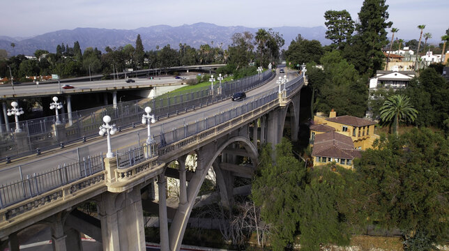 Aerial view of the historic Colorado Street Bridge, a Beaux-Arts concrete arch bridge with ornate lamps, surrounded by trees and highway infrastructure in Pasadena, California, United States.