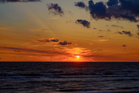 Dramatic sunset over a dark, rolling sea with glowing orange skies, scattered clouds, and the sun setting on the horizon.