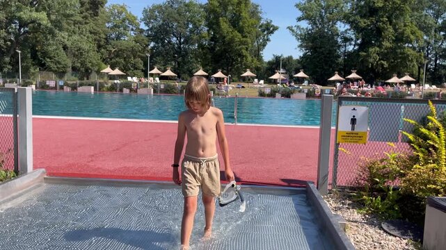 Boy walking through shallow pool area outdoors towards green trees, active outdoor lifestyle, summer leisure concept, summer holiday spirit.