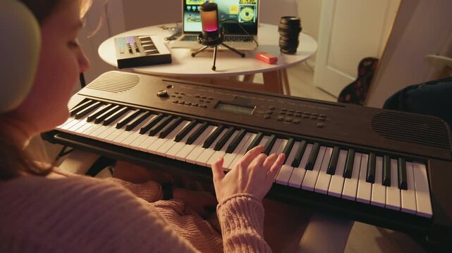 Person seated at electronic keyboard playing with both hands, fingers moving across keys in warm home living room with lamp, phone stand, mug, notebook and relaxed practice atmosphere