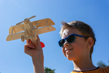 A boy in glasses holds a children's wooden airplane in his hand against the background of the blue sky in the park. © Prrrettty