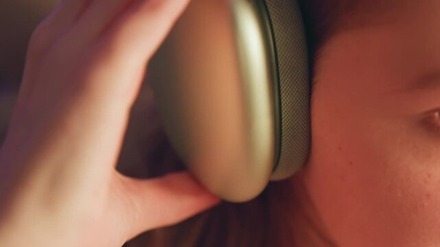 Warm indoor side-profile scene of young woman adjusting padded over-ear headphones by hand, with calm focused expression, visible eye detail, soft light, and subtle personal listening moment